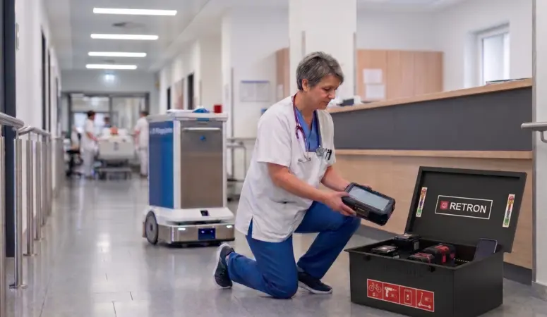 Eine Pflegekraft kniet in einem Krankenhausflur und nimmt einen Akku aus einer RETRON BOX. Im Hintergrund ist ein autonomer Transportroboter auf dem Flur zu sehen.